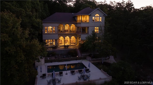 back of house at dusk featuring a balcony, an outdoor pool, a patio, stairs, and stucco siding