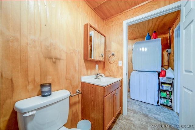 half bath featuring toilet, wood walls, stacked washer / drying machine, and vanity