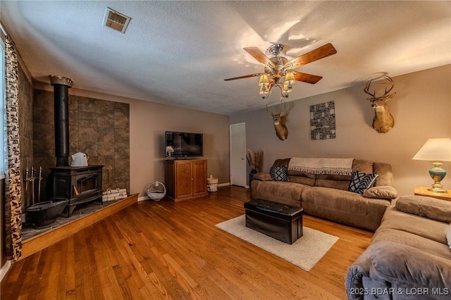 living area featuring visible vents, a wood stove, a ceiling fan, a textured ceiling, and wood finished floors