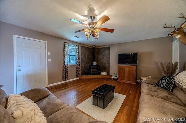 living room featuring baseboards, a ceiling fan, a wood stove, visible vents, and wood finished floors