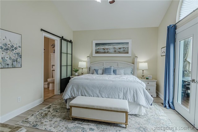 bedroom with a barn door, vaulted ceiling, light wood finished floors, ensuite bath, and baseboards