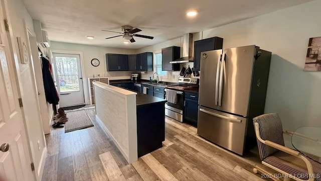 kitchen featuring a sink, wall chimney range hood, ceiling fan, appliances with stainless steel finishes, and light wood-style floors