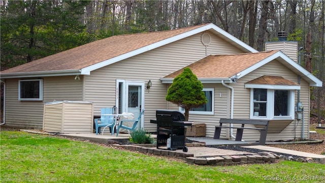 back of property with a yard, a shingled roof, a patio, and a chimney