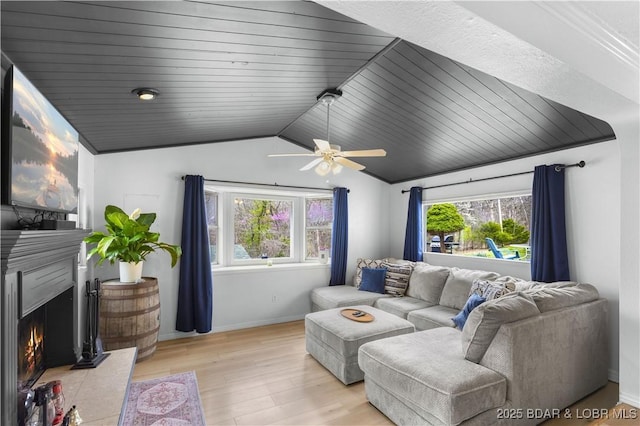 living area featuring baseboards, ceiling fan, light wood-type flooring, vaulted ceiling, and a lit fireplace