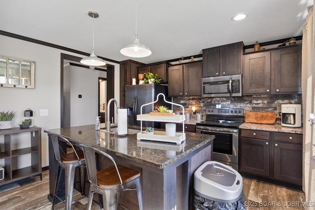 kitchen with appliances with stainless steel finishes, dark brown cabinetry, a kitchen island with sink, decorative backsplash, and wood finished floors