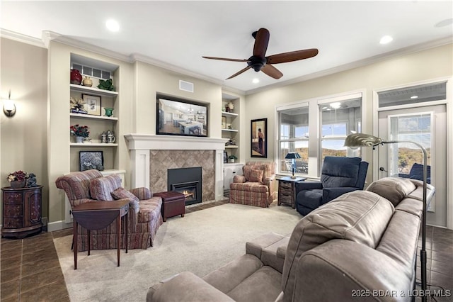 living room featuring a tiled fireplace, built in shelves, crown molding, a ceiling fan, and visible vents