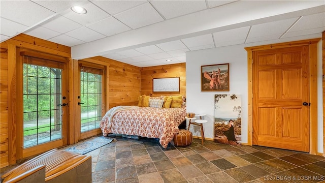 bedroom with wooden walls, access to outside, baseboards, stone finish flooring, and a drop ceiling