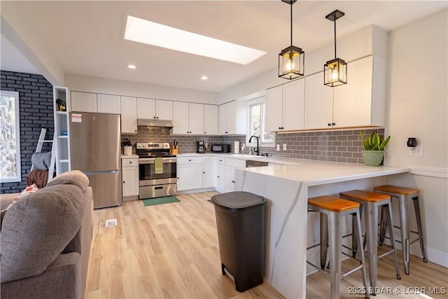 kitchen with light countertops, light wood finished floors, a peninsula, under cabinet range hood, and stainless steel appliances