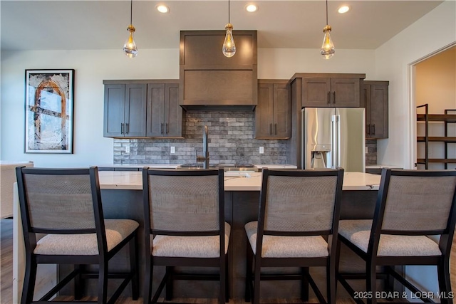 kitchen featuring stainless steel refrigerator with ice dispenser, light countertops, a kitchen island with sink, and tasteful backsplash