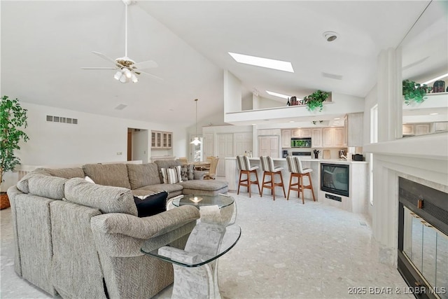 living area with ceiling fan, visible vents, a skylight, high vaulted ceiling, and a glass covered fireplace