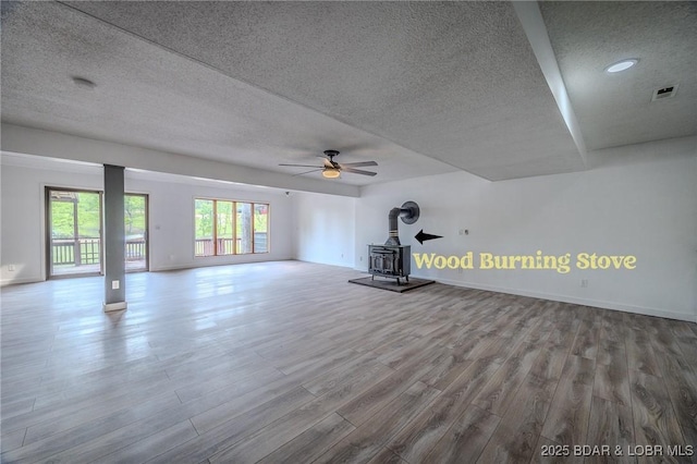 unfurnished living room with a wood stove, a textured ceiling, a ceiling fan, wood finished floors, and visible vents