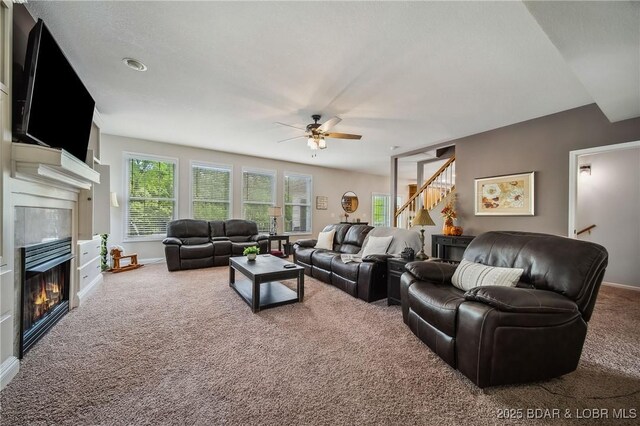 carpeted living area featuring baseboards, a ceiling fan, a glass covered fireplace, and stairway