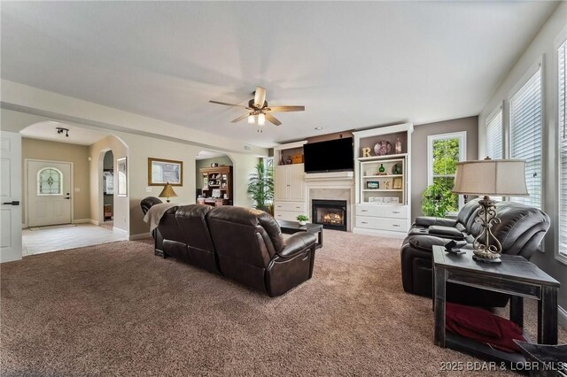 carpeted living room featuring arched walkways, a ceiling fan, a glass covered fireplace, and a wealth of natural light