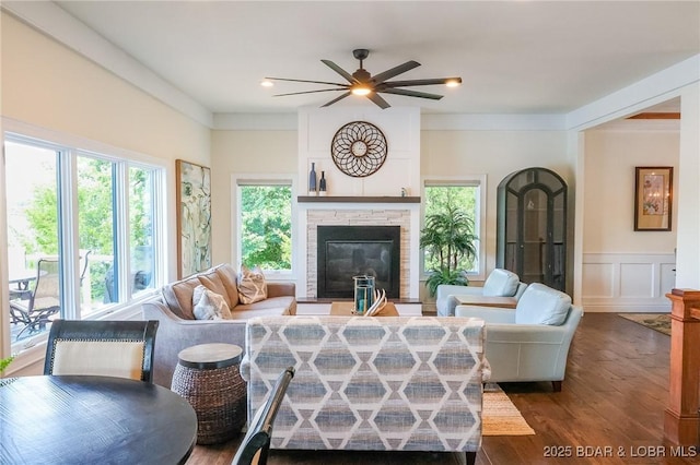 living room featuring a decorative wall, a fireplace, dark wood-type flooring, a wainscoted wall, and a ceiling fan