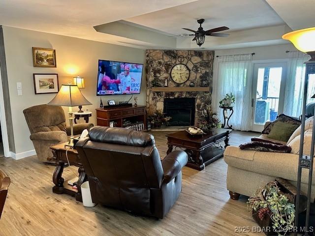 living room featuring a ceiling fan, a stone fireplace, wood finished floors, a raised ceiling, and baseboards