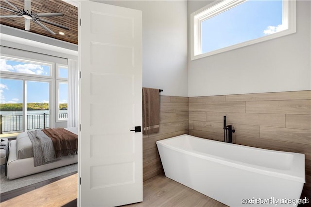 ensuite bathroom with tile walls, wood finished floors, a ceiling fan, a freestanding bath, and a water view