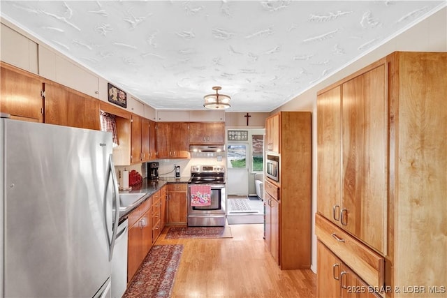 kitchen with appliances with stainless steel finishes, brown cabinetry, light wood-style flooring, under cabinet range hood, and a chandelier