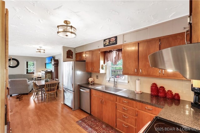 kitchen with appliances with stainless steel finishes, a sink, ventilation hood, dark countertops, and light wood-type flooring