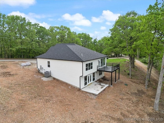 view of property exterior with cooling unit, a patio, a deck, a shingled roof, and view of scattered trees