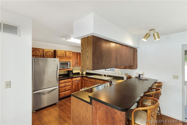 kitchen featuring stainless steel appliances, a peninsula, dark countertops, a breakfast bar, and brown cabinetry
