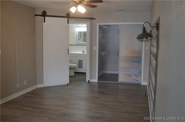 unfurnished bedroom with wood finished floors, ensuite bathroom, baseboards, a barn door, and ceiling fan