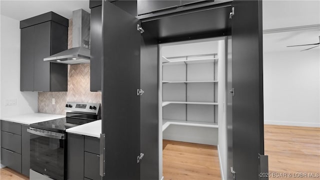 kitchen featuring stainless steel electric range, wall chimney range hood, light wood-type flooring, decorative backsplash, and light countertops
