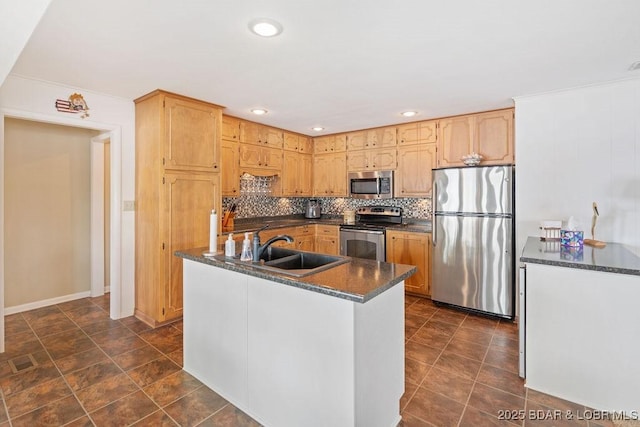 kitchen with stainless steel appliances, a sink, dark countertops, decorative backsplash, and an island with sink
