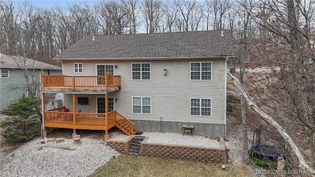 back of property with a trampoline, a deck, a shingled roof, and a patio