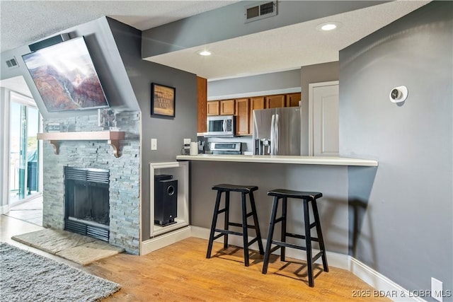 kitchen with stainless steel appliances, brown cabinetry, light wood-style flooring, a breakfast bar area, and a stone fireplace