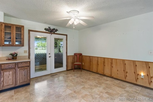 doorway to outside featuring french doors, a wainscoted wall, a textured ceiling, ceiling fan, and wooden walls