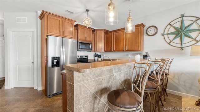 kitchen featuring stainless steel appliances, brown cabinetry, a peninsula, a breakfast bar area, and hanging light fixtures