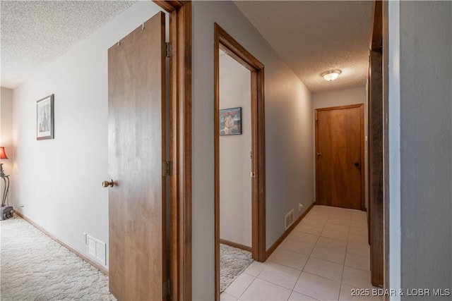 hallway with a textured ceiling, light tile patterned floors, baseboards, and light colored carpet