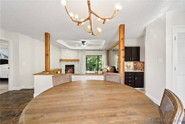 unfurnished dining area featuring ceiling fan, a stone fireplace, a textured ceiling, a chandelier, and a raised ceiling