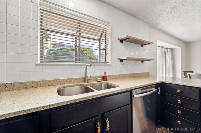 kitchen with stainless steel dishwasher, a sink, dark cabinetry, backsplash, and light stone counters