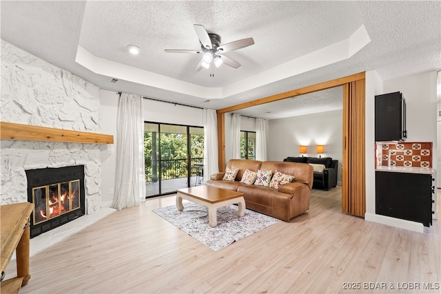 living room with a textured ceiling, a tray ceiling, a ceiling fan, a stone fireplace, and light wood-style flooring