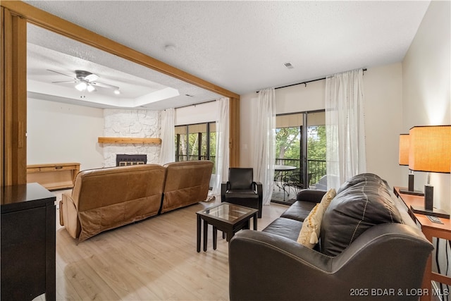 living area with light wood-style floors, a textured ceiling, a stone fireplace, a ceiling fan, and a tray ceiling