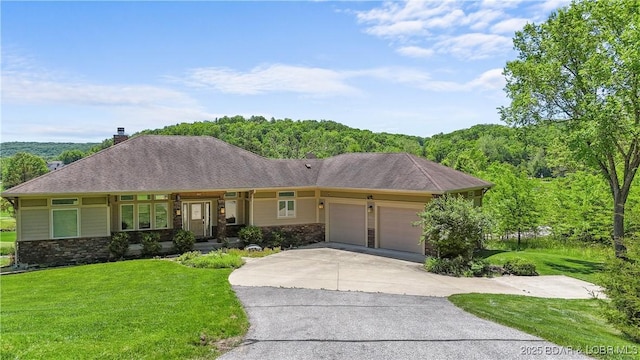 ranch-style house with a garage, a front lawn, driveway, a chimney, and a wooded view