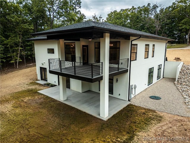 back of house featuring a balcony, a patio area, stucco siding, a shingled roof, and view of wooded area