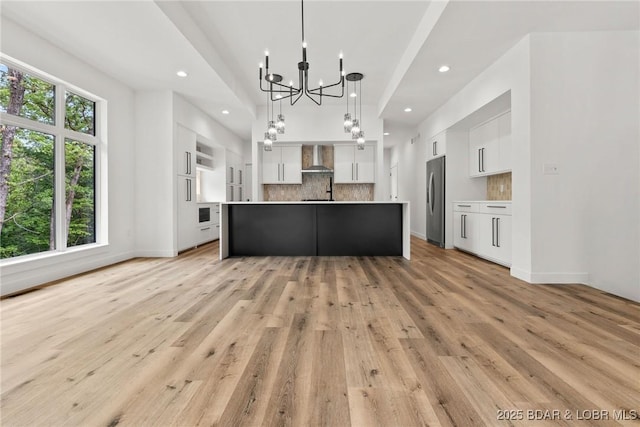 kitchen with freestanding refrigerator, wall chimney range hood, a chandelier, white cabinetry, and backsplash