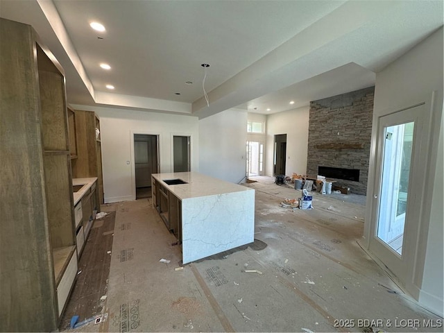 kitchen featuring a stone fireplace, a large island with sink, brown cabinetry, recessed lighting, and a raised ceiling