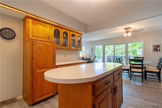 kitchen featuring brown cabinetry, light countertops, a kitchen island, glass insert cabinets, and light wood-style floors
