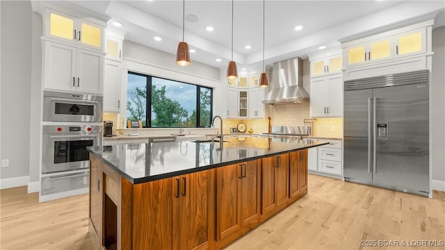 1254 Darwin Dr, Osage Beach, MO 65065 - kitchen with appliances with stainless steel finishes, wall chimney range hood, a warming drawer, light wood finished floors, and a center island with sink