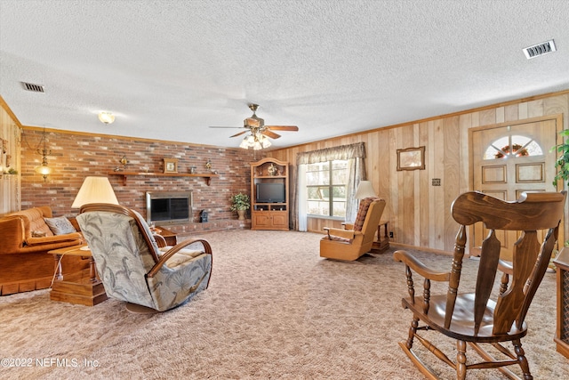 carpeted living room featuring a brick fireplace, ceiling fan, crown molding, wooden walls, and a textured ceiling