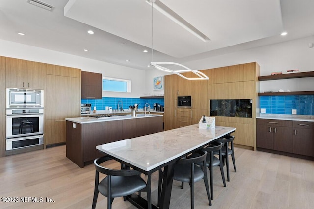 kitchen featuring appliances with stainless steel finishes, backsplash, a kitchen island with sink, pendant lighting, and light hardwood / wood-style flooring