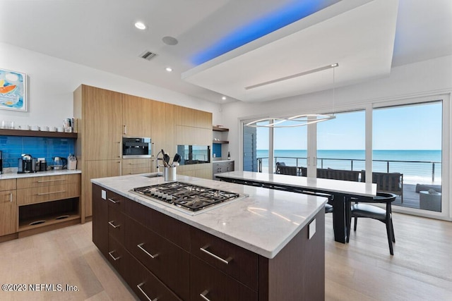 kitchen with wall oven, stainless steel gas stovetop, a center island with sink, a water view, and light wood-type flooring