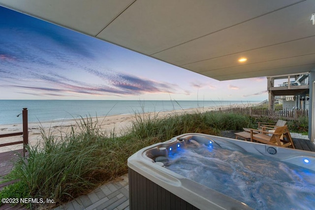 pool at dusk with a water view, a beach view, and a hot tub