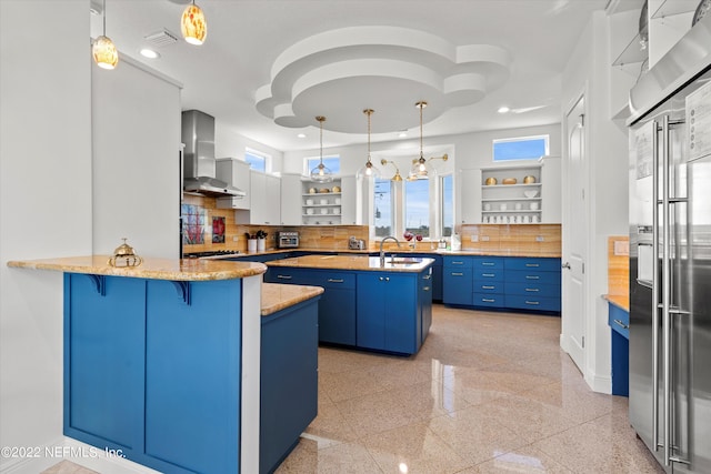 kitchen with wall chimney range hood, blue cabinets, backsplash, kitchen peninsula, and white cabinets