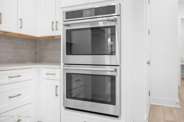 kitchen with stainless steel double oven, white cabinets, light wood-type flooring, and tasteful backsplash