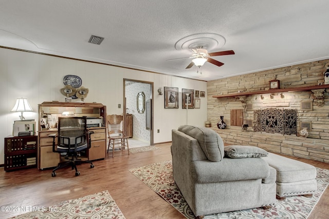 living room featuring a stone fireplace, a textured ceiling, ornamental molding, ceiling fan, and hardwood / wood-style floors