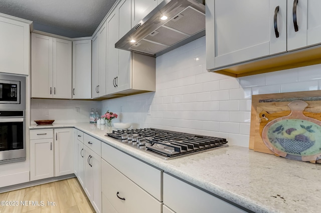 kitchen featuring backsplash, wall chimney range hood, light hardwood / wood-style flooring, light stone countertops, and stainless steel appliances